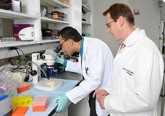 A man in a white coat examines an item through a microscope through the MSTP program, while another man in a white coat stands behind him.